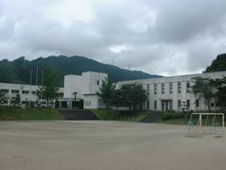緑の山をバックに広い校庭がある白い外壁の匹見小学校の外観の写真