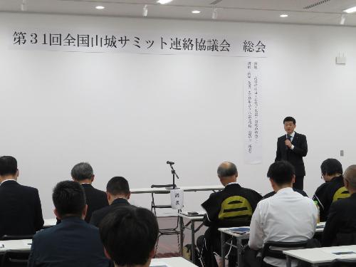グラントワ多目的ホールで総会参加者らの前で挨拶をしている市長の写真