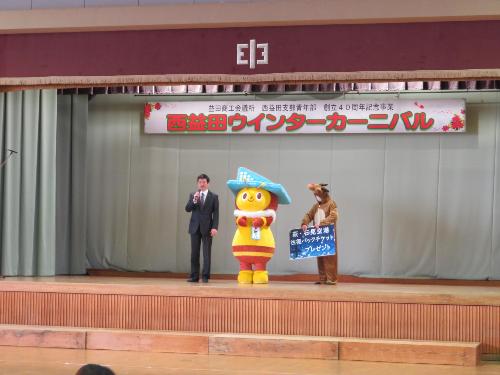 横田中学校体育館ステージ上で空港キャラクターのトビーくんと一緒に立ち参加者らに挨拶をしている市長の写真