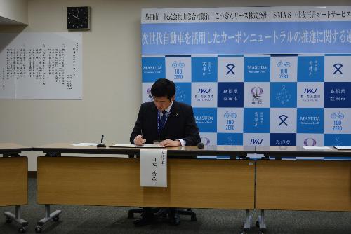 市役所大会議室で机に向かい協定書にサインをしている市長の写真