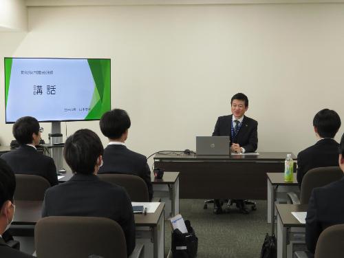市役所大会議室で新規採用職員らにモニターを使い講話を行っている市長の写真