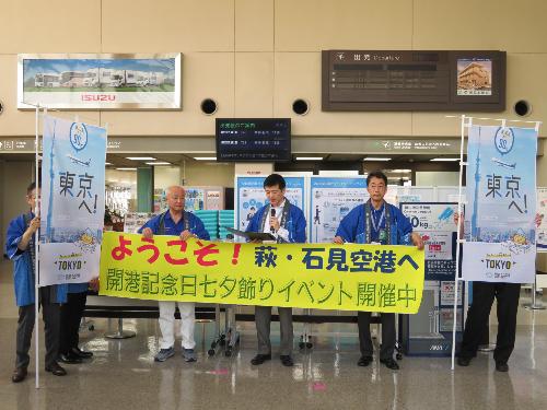 萩・石見空港出発ロビーで歓迎の垂れ幕を持ち搭乗者らに挨拶をしている市長の写真