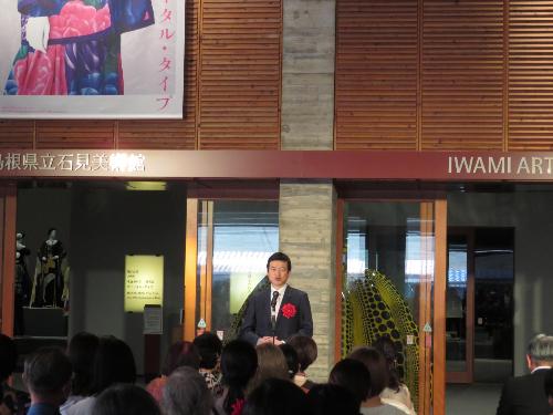 グラントワ企画展示室前のロビーで内覧会出席者らに祝辞を述べている市長の写真