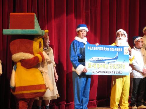グラントワステージ上で空港キャラクターのトビーくんらと一緒に抽選商品の空港往復券について説明している市長の写真