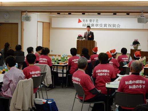 光永榮子氏緑綬褒章受章報告会にて挨拶を述べる市長と耳を傾ける参加者らの写真