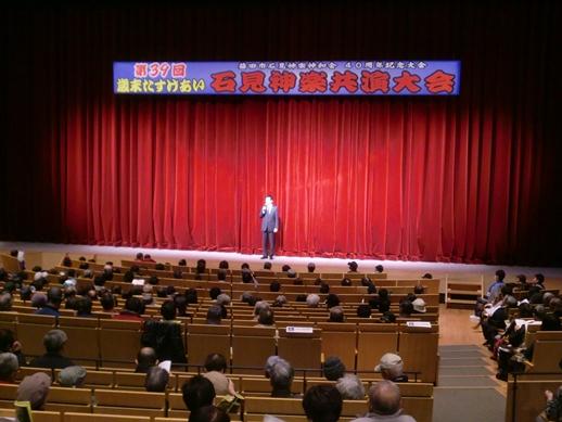 歳末たすけあい石見神楽共演大会のステージで挨拶を述べる市長と観客らの写真