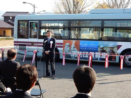 消防団募集広告ラッピングバスと紅白リボンテープの前に立ち出発式の挨拶を述べる市長の写真