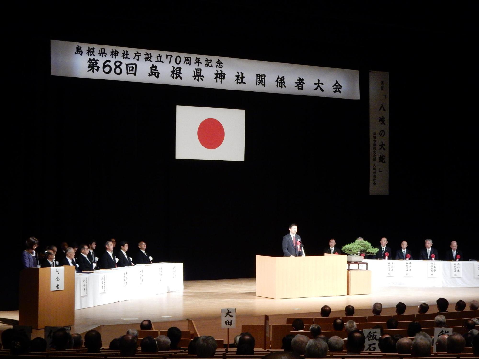 島根県神社関係者大会のステージで挨拶を述べる市長と傾聴する出席者らの写真