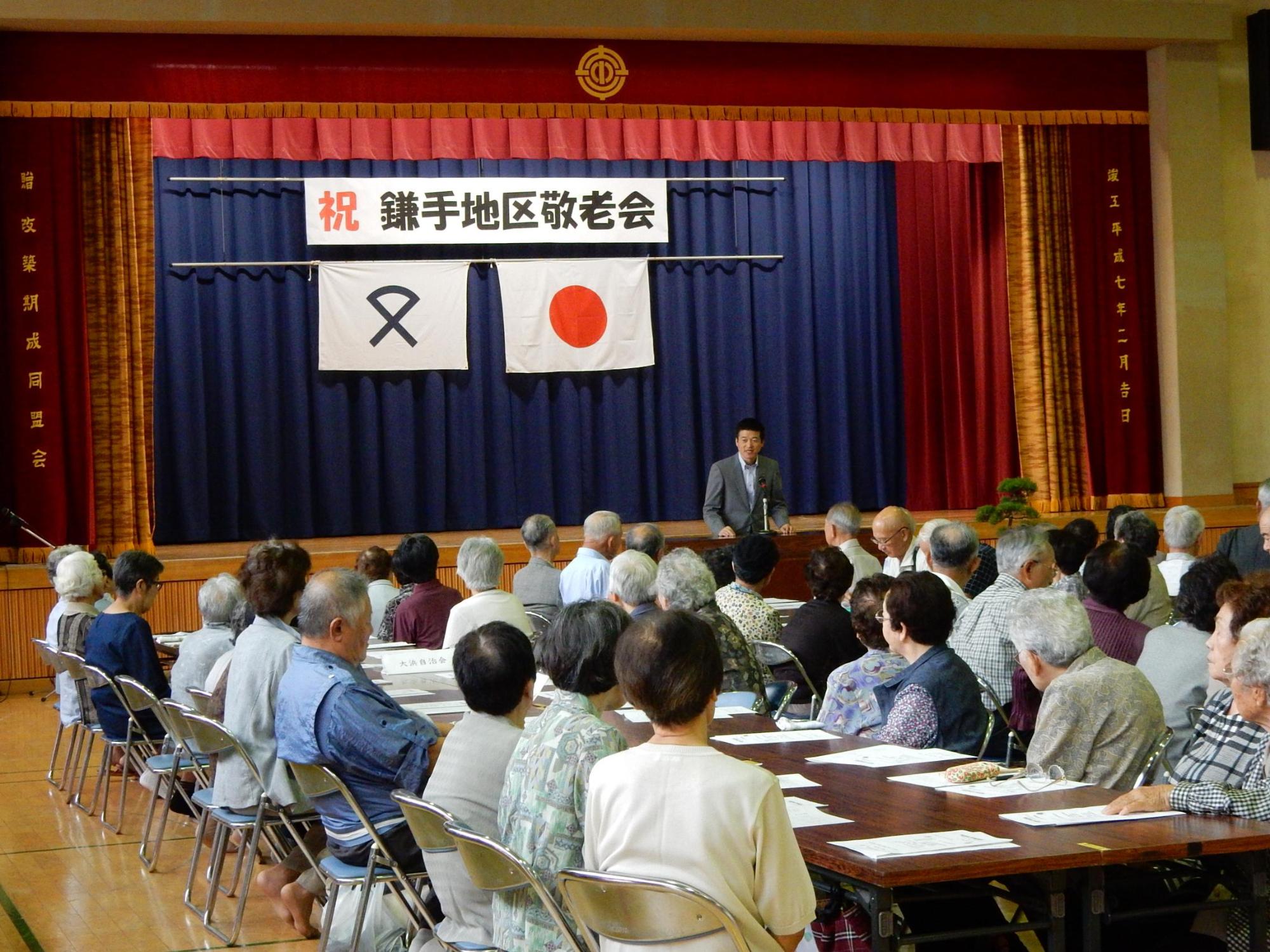 鎌手地区敬老会にて挨拶を述べる市長と傾聴する出席者らの写真