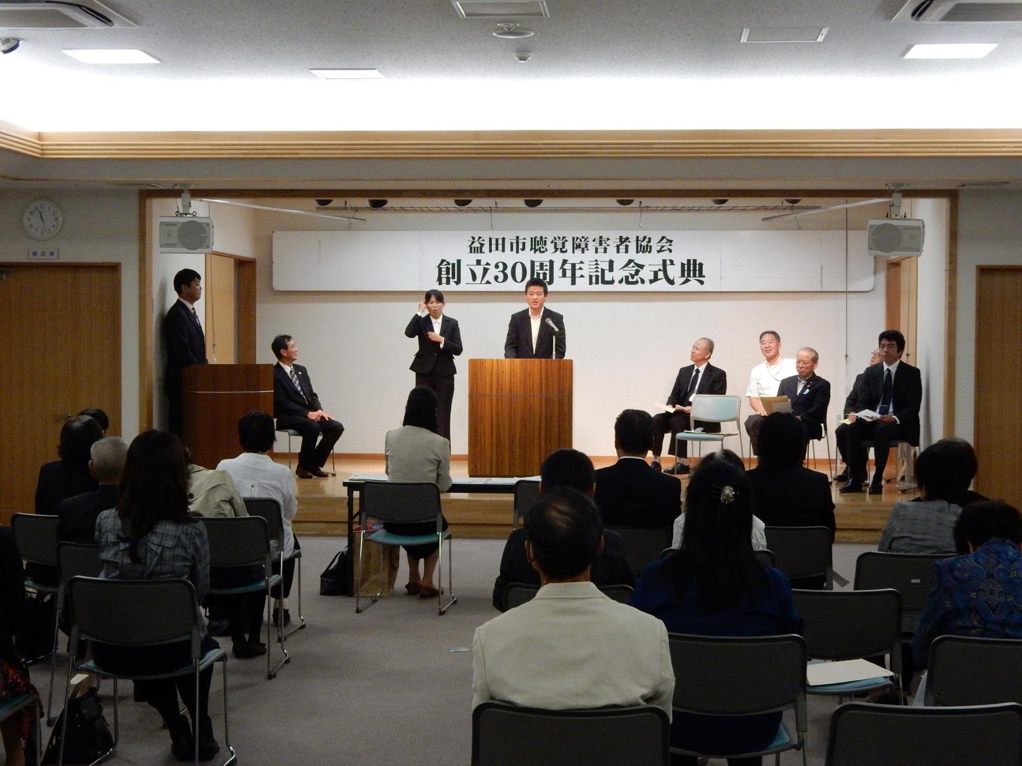 益田市聴覚障害者協会記念式典の壇上で手話通訳士と共に挨拶を述べる市長の写真