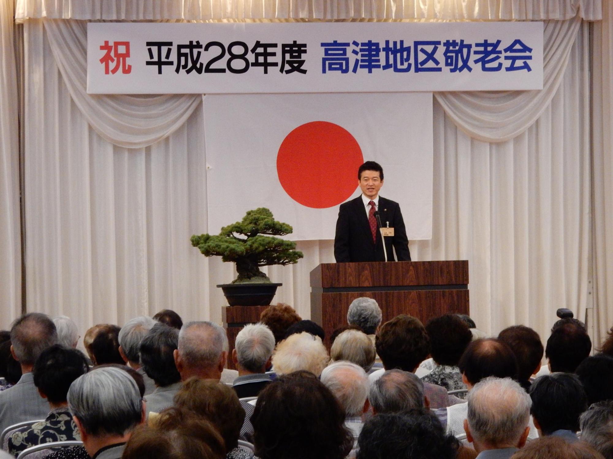 高津地区敬老会にて挨拶を述べる市長と傾聴する出席者らの写真