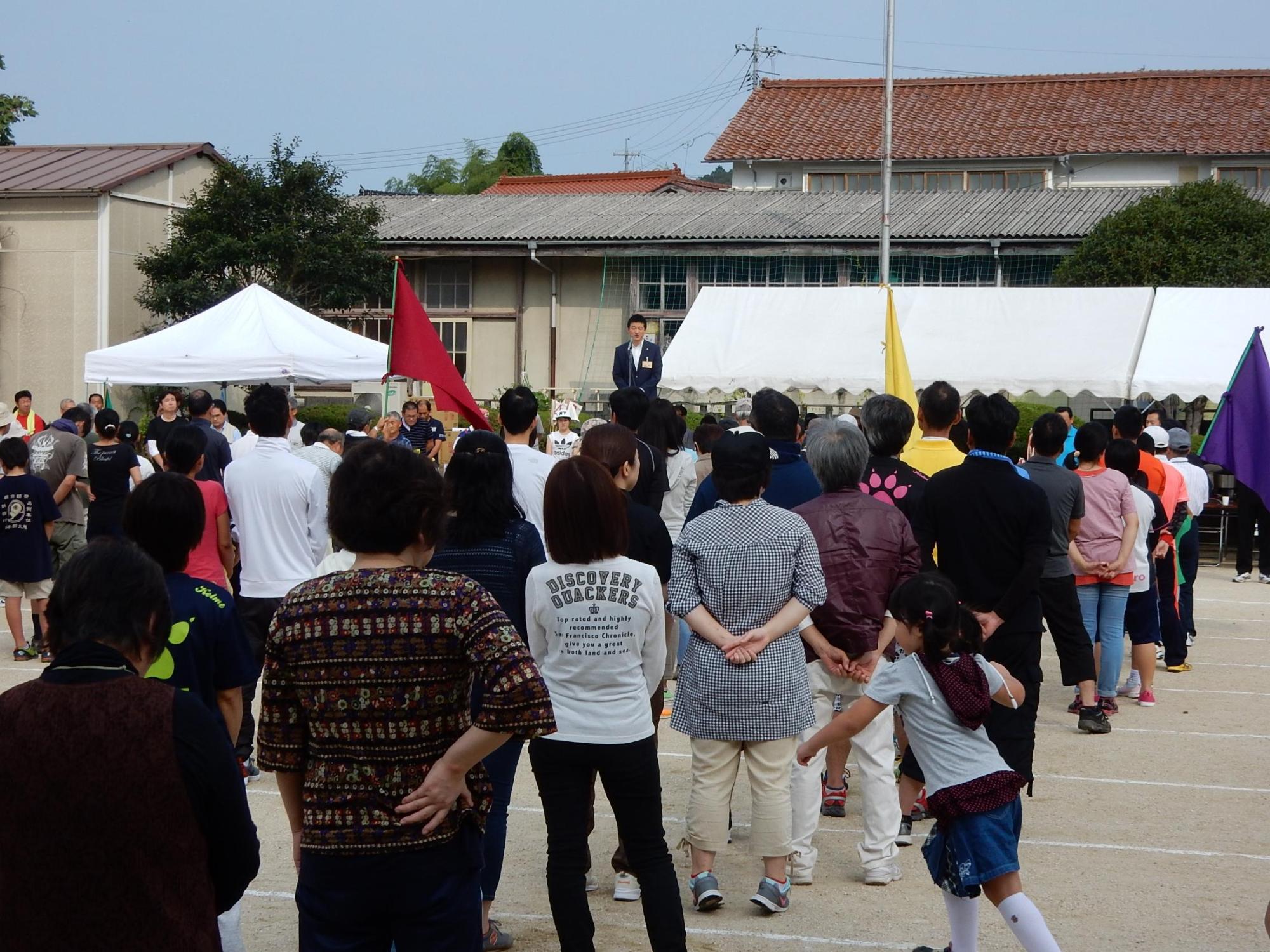中西地区民体育大会にてグランドのステージで挨拶を述べる市長と傾聴する参加者らの写真