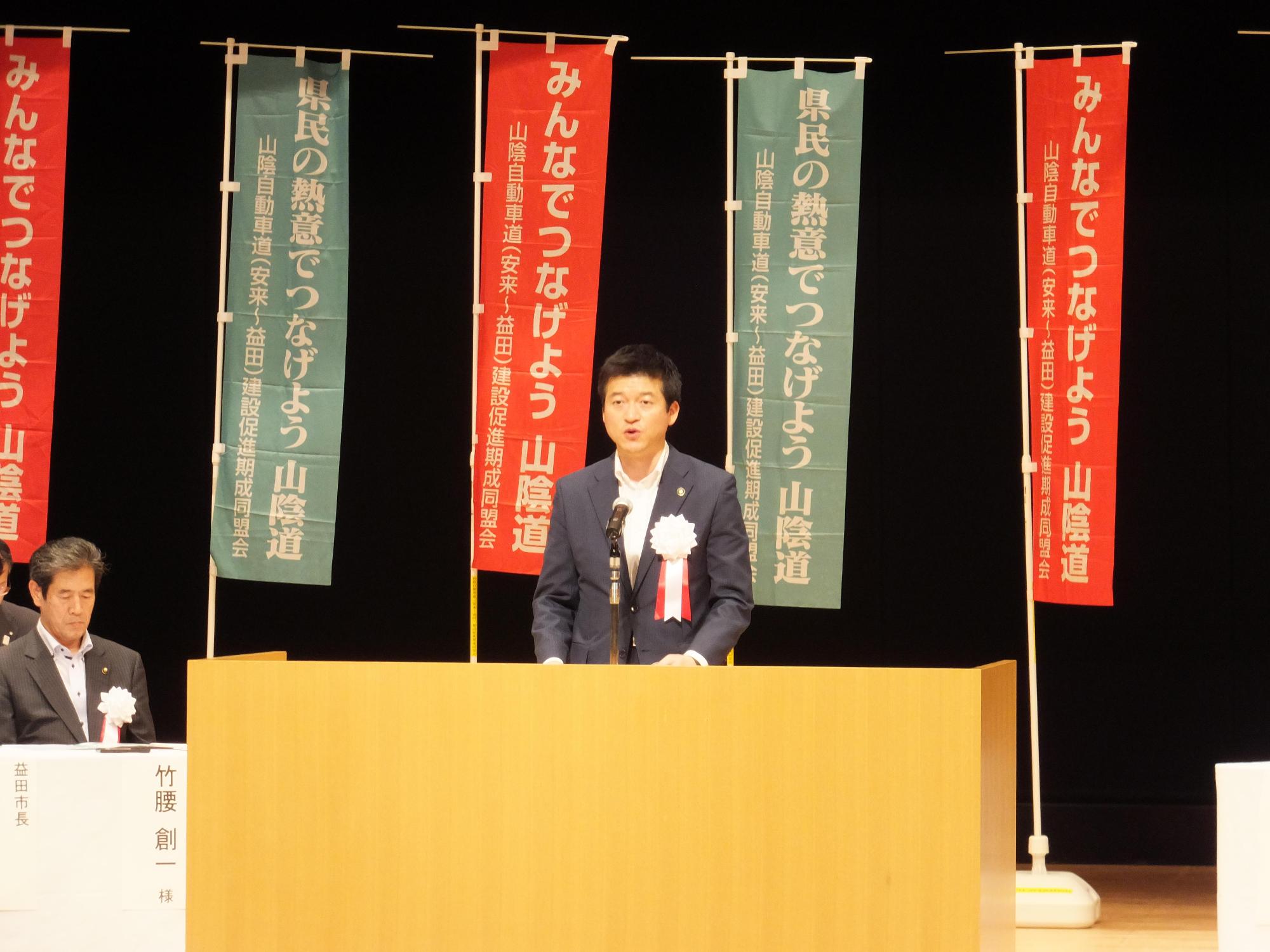 背後にのぼりが立ち並ぶ山陰道総決起大会会場ステージ前で言葉を述べる市長の写真
